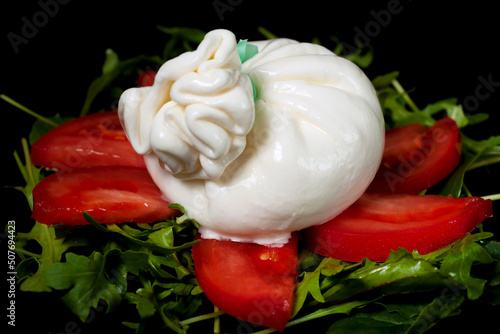 Italian burrata cheese on a salad of arugula greens and sliced tomatoes on a black background.