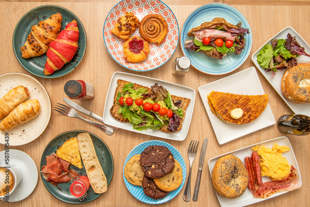 Overhead view of a set of dessert plates, cinnamon roll, cookies, assorted croissants, lettuce sandwiches, chocolate chips, and egg breakfasts