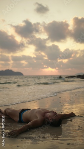 The young handsome man spends his vacation on a tropical island. Concept relaxation meditation tourism, wellness health body. The guy laying down on the wet sand on beach. Evening dusk view