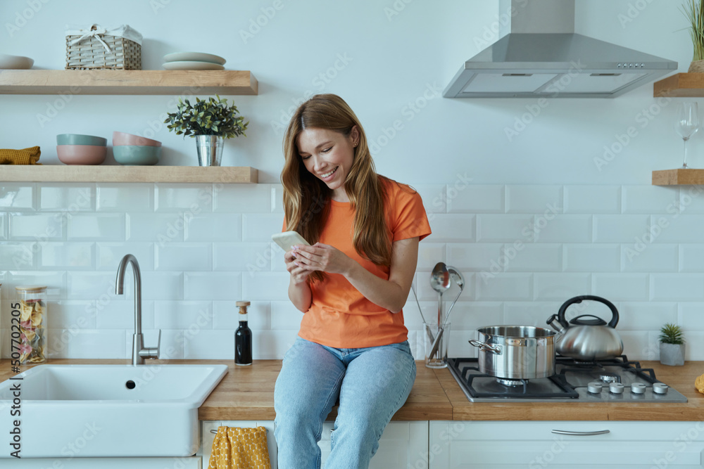 Beautiful young woman using smart phone while sitting at the kitchen counter Beautiful young woman using smart phone while sitting at the kitchen counter