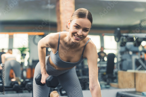 Fit woman exercising with dumbbell weights on the bench at the gym and smiling at the camera during daytime. High quality photo