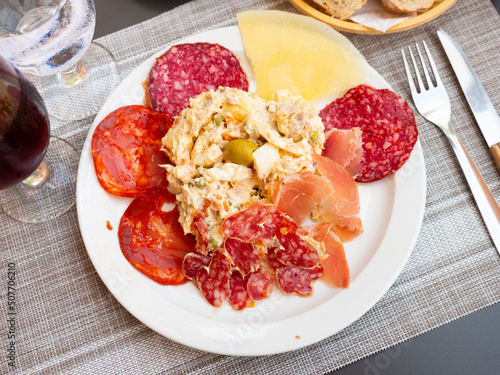 Plate with coldcuts from sliced salami, fuet, chorizo, jamon iberico and hard cheese served with russain salad. Popular Spanish entremets photo