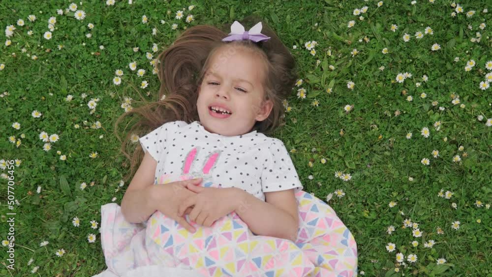 Little happy child girl in dress with bow on head laying on green lawn in the park. Summer time, nature, Dreams, Lifestyle Concepts. Smilling Baby Face close up. emotional child grimaces at camera Little happy child girl in dress with bow on head laying on green lawn in the park. Summer time, nature, Dreams, Lifestyle Concepts. Smilling Baby Face close up. emotional child grimaces at camera