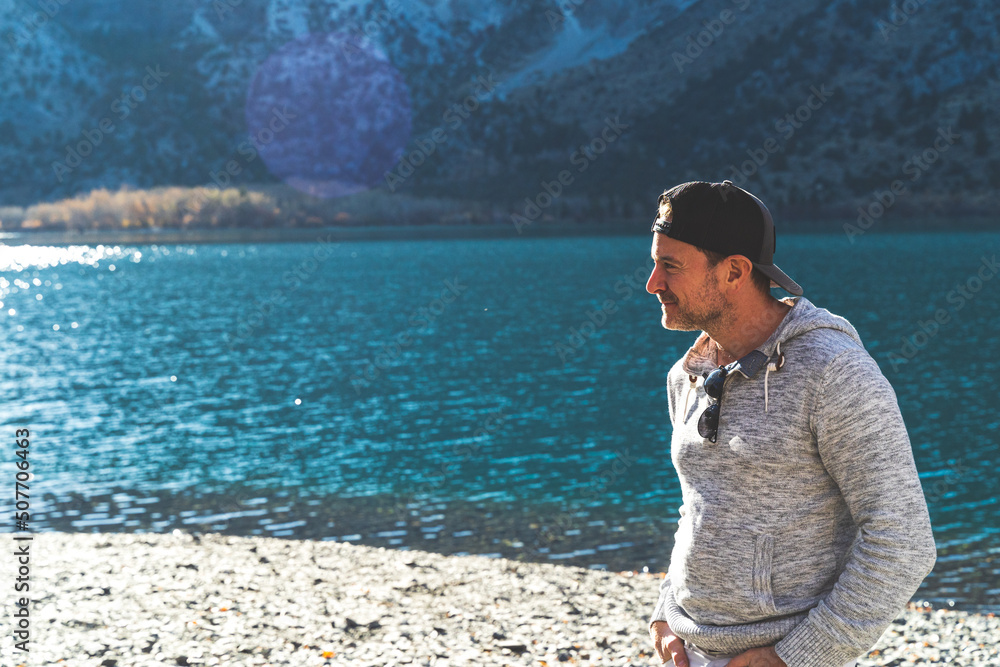 Man standing in front of mountain lake with ball cap on looking away Man standing in front of mountain lake with ball cap on looking away