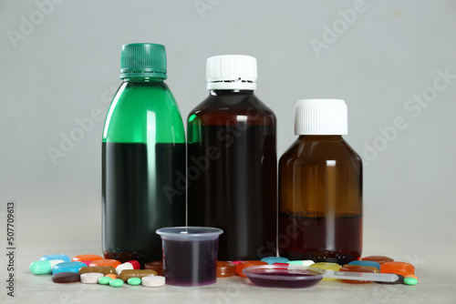 Bottles of cough syrup, dosing spoon, measuring cup and pills on light background