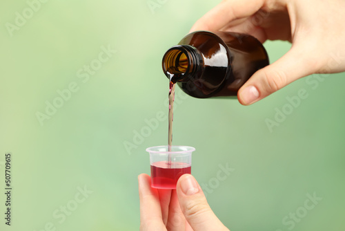 Woman pouring cough syrup into measuring cup on light green background, closeup
