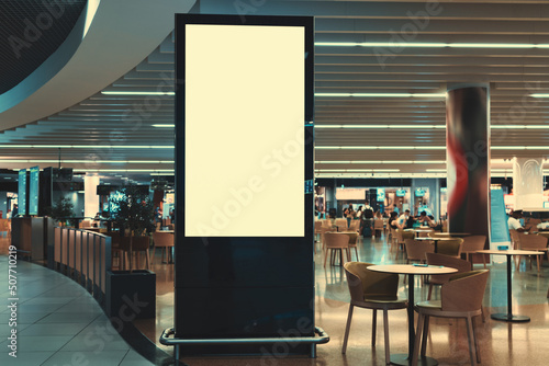 A blank vertical advertising billboard in front of a crowded mall food court with tables and customers in the background. An empty LCD screen template for copy space in front of an airport restaurant