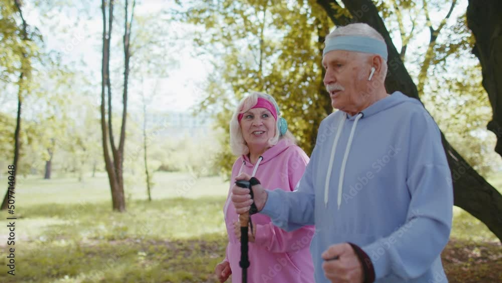 Cheerful senior elderly caucasian couple jogger grandparents training Nordic walking with ski trekking poles, running in summer park outdoor. Mature grandfather, grandmother hiking leisure sport hobby Cheerful senior elderly caucasian couple jogger grandparents training Nordic walking with ski trekking poles, running in summer park outdoor. Mature grandfather, grandmother hiking leisure sport hobby