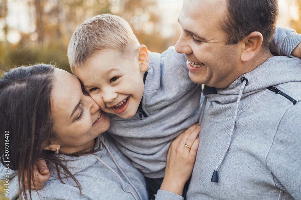 Fototapeta premium Happy parents and son are smiling together while standing in the park, posing in the daytime. The perfect little family. Family in nature.