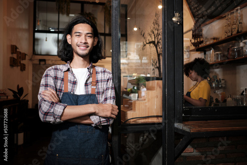 A long hair handsome Asian male startup barista with apron standing at casual cafe door, arms crossed, looking at the camera with and welcoming smile, happy and cheerful with coffee shop service jobs. © tigercat_lpg
