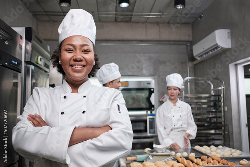 Foto Young African American female chef in white cooking uniform looks at camera, arms crossed and cheerful smile with food professional occupation, commercial pastry culinary jobs in a restaurant kitchen