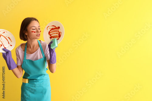 Young woman with cleaning sponge and dirty dishes on yellow background