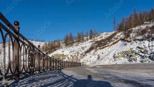 A metal fence is installed on the side of the road. An openwork shadow on the asphalt. A mountain with bare trees on snow-covered slopes against a blue sky. Altai