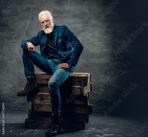 Photo of handsome aged man with long beard and stylish hairstyle sitting on firewoods.