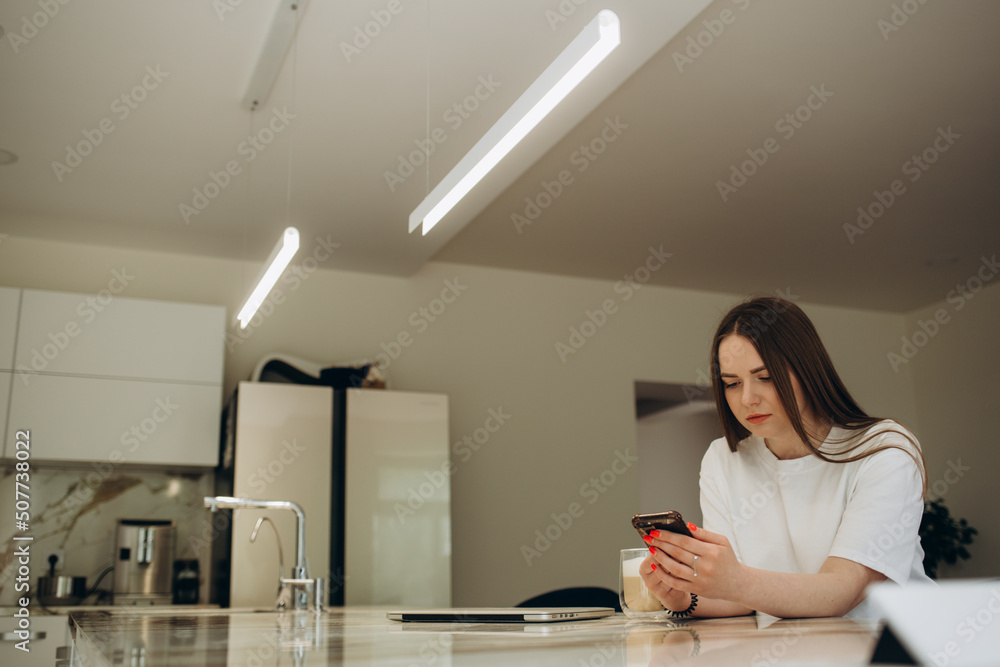 Young woman using smartphone leaning at kitchen table with coffee mug and organizer in a modern home. Smiling woman reading phone message. Brunette happy girl typing a text message Young woman using smartphone leaning at kitchen table with coffee mug and organizer in a modern home. Smiling woman reading phone message. Brunette happy girl typing a text message