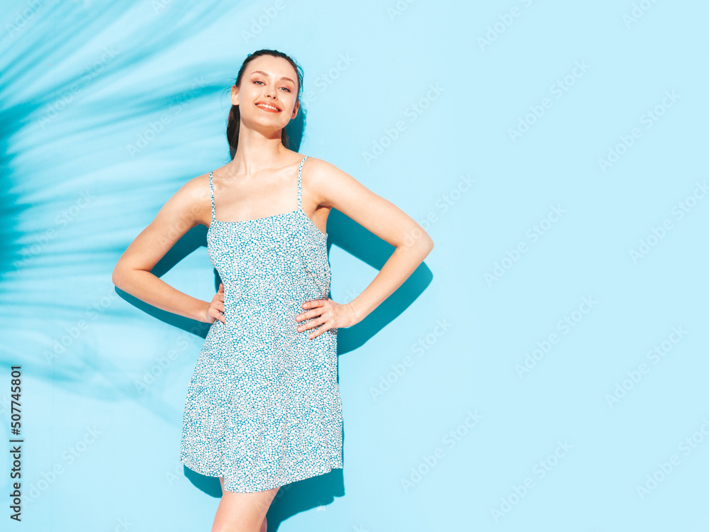 Fototapeta premium Young beautiful smiling female in trendy summer dress. Sexy carefree woman posing near blue wall in studio. Positive brunette model having fun.Cheerful and happy. Isolated