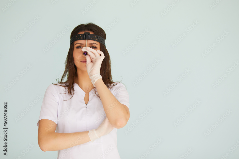 Cute young female otolaryngologist standing with illuminator on her head on bright background Cute young female otolaryngologist standing with illuminator on her head on bright background