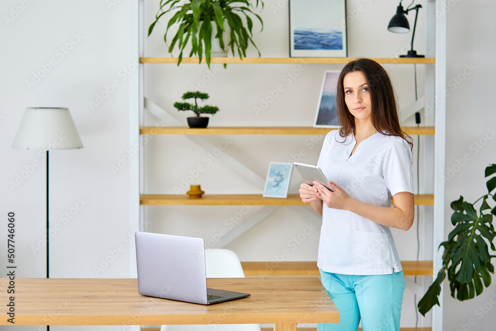 Smiling female doctor wearing uniform using tablet, standing in office, browsing medical apps, professional therapist physician working online, chatting, consulting patient Smiling female doctor wearing uniform using tablet, standing in office, browsing medical apps, professional therapist physician working online, chatting, consulting patient