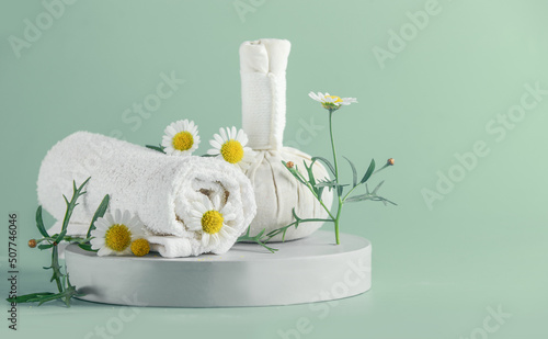 Wellness setting with folded white towel, chamomile flowers and herbal massage stamp on product podium with petals at pastel turquoise background. Front view.