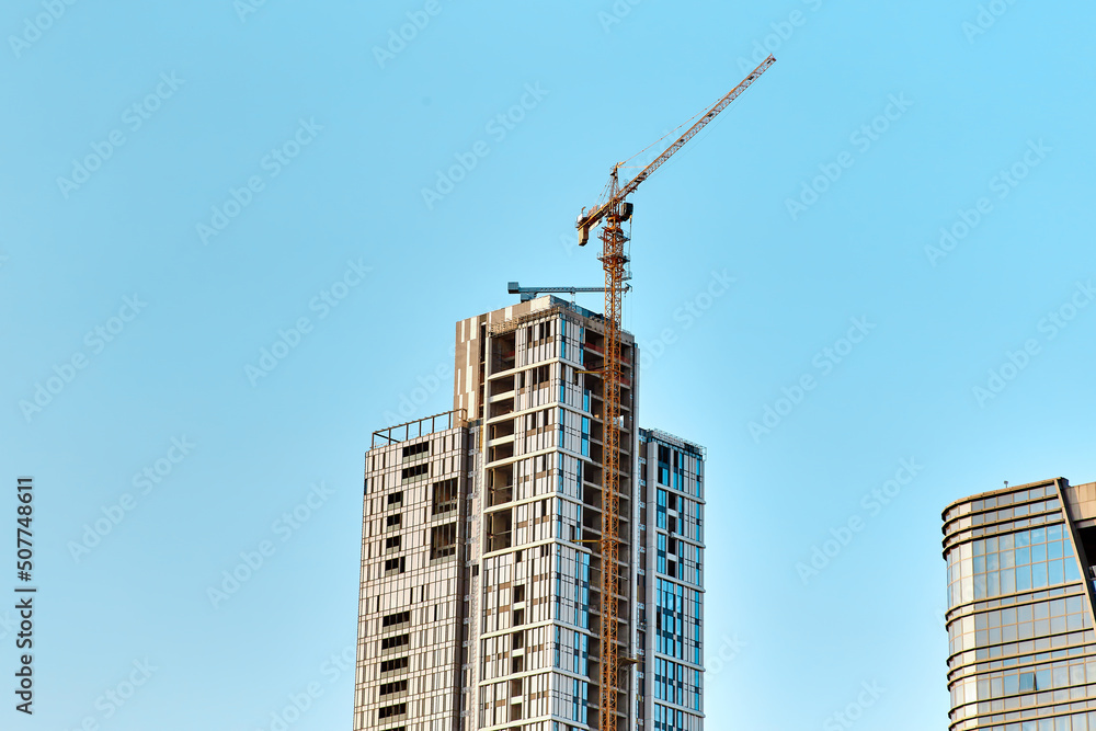 Top of the yellow tower crane on the construction of white with glass skyscraper near another modern building against a blue sky with smaller crane at the bottom in sunny weather Top of the yellow tower crane on the construction of white with glass skyscraper near another modern building against a blue sky with smaller crane at the bottom in sunny weather