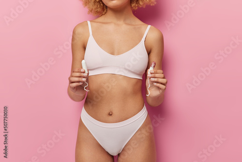 Woman body with tampons for menstruation dressed in white underwear isolated over pink background. Faceless female model holds intimate hygiene products during periods. Protective care concept