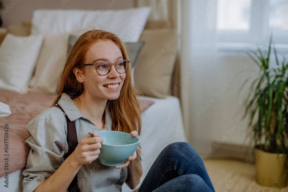 Happy woman with bowl of food sitting in front of bed at home Happy woman with bowl of food sitting in front of bed at home