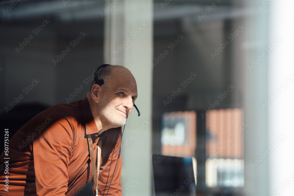 Smiling businessman wearing headset in office seen through window Smiling businessman wearing headset in office seen through window