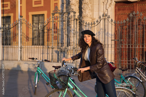 Young and beautiful woman, brunette, with curly hair, with leather jacket and black beret, riding a bicycle while visiting the city. Concept beauty, fashion, trend, travel, holidays, guided tours. photo