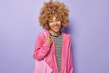 Indoor shot of curly haired European woman smiles pleasantly wears casual striped jumper and pink jacket carries fabric bag being in good mood isolated over purple background. Happy emotions