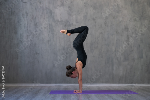 Side view portrait beautiful young woman wearing black sportswear, exercising against a gray wall doing yoga or pilates. Standing in a pose. Full length