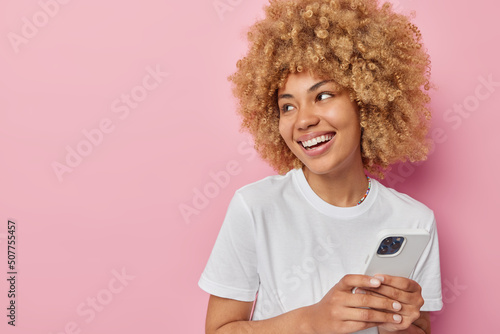 Smiling beautiful woman with curly bushy hair uses mobile phone chats online in messanger dressed in casual white t shirt focused away isolated over pink background blank space for your promotion photo