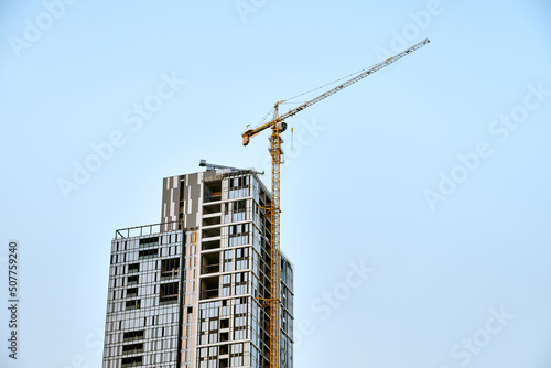 Top of the yellow tower crane on the construction of white with glass modern skyscraper against a blue sky with smaller crane at the bottom