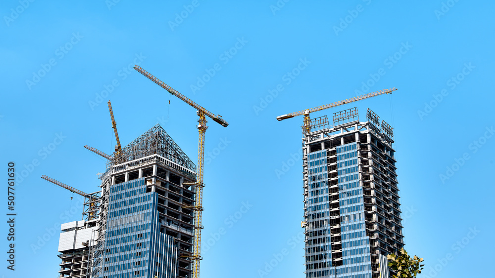 Construction of two modern skyscrapers with glasses with yellow tower cranes along the buildings witn another buildings under construction around in bright sunny weather against a clear blue sky