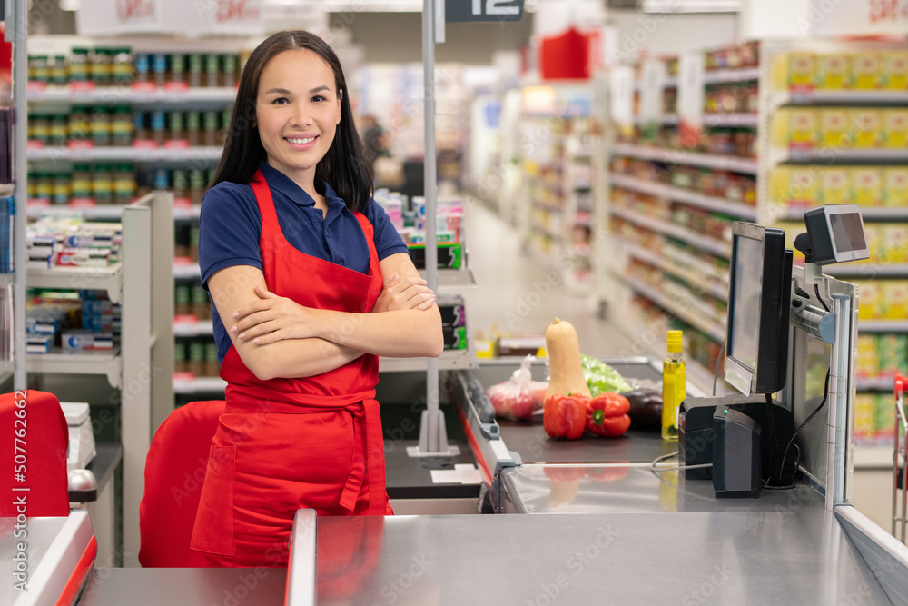 Attractive Asian woman working in supermarket standing at cash desk with arms crossed looking at camera smiling Attractive Asian woman working in supermarket standing at cash desk with arms crossed looking at camera smiling
