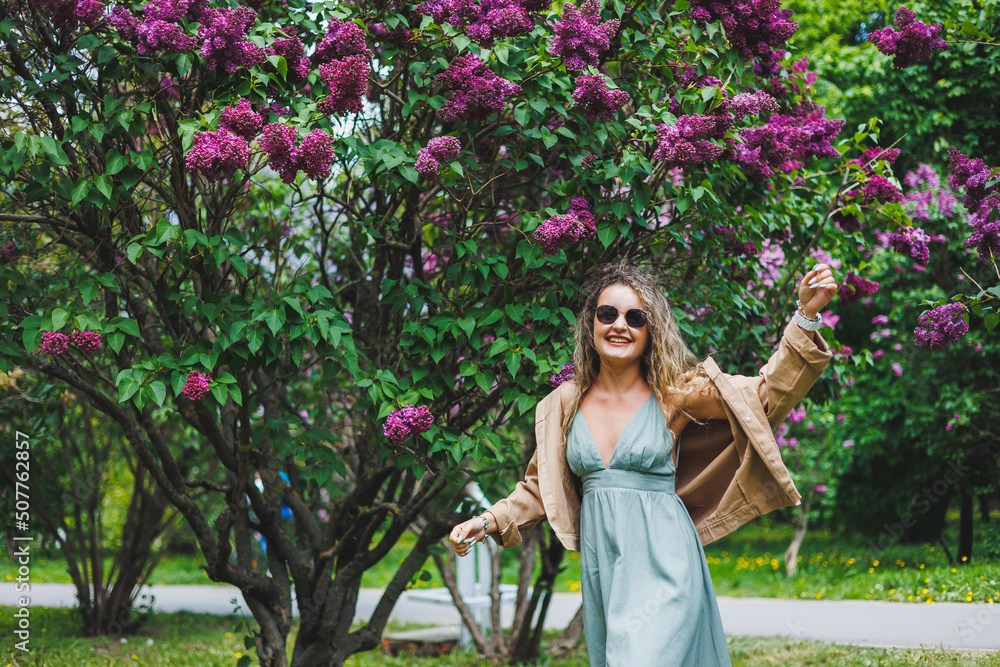 Naklejka premium A beautiful curly woman in a summer dress runs and rejoices, she is wearing sunglasses. Against the background of a purple lilac bush