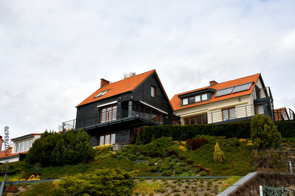 A view of a white and a black house standing on the top of a tall hill covered with shrubs, grass, and other flora spotted during a hike on a cloudy yet warm spring day in Poland