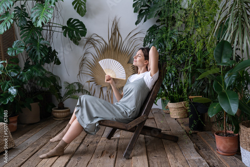 Happy young caucasian woman florist relax in home garden breathing fresh air hold paper fan in hand. Relaxed carefree millennial female gardener refreshing in indoor orangery during hot summer day