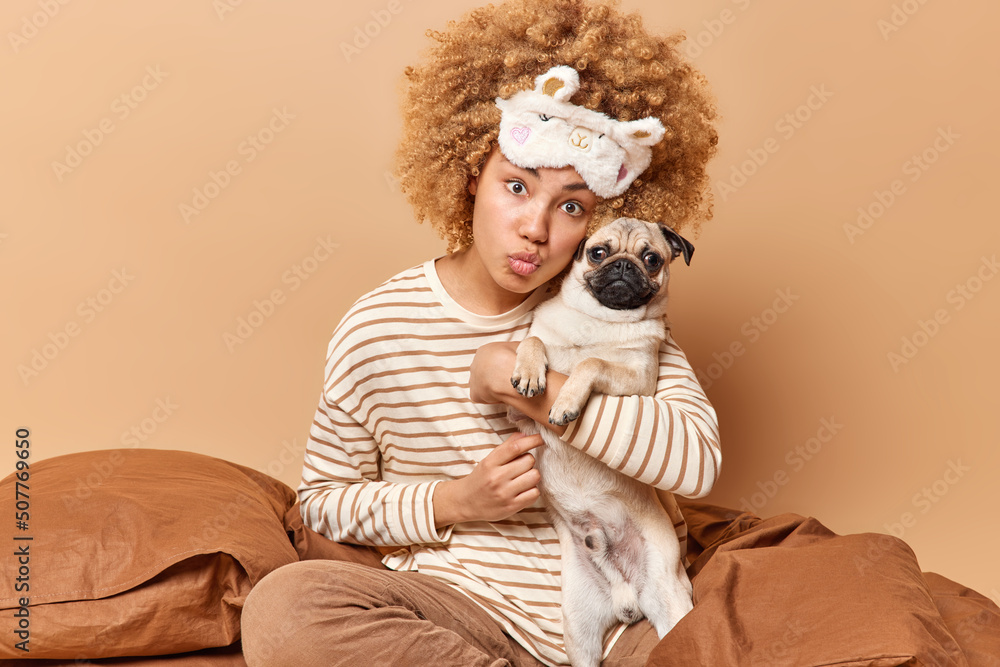 Indoor shot of lovely woman dresssed in pajama embraces favorite pet with love sits in bed keeps lips folded has romantic expression wears sleepmask isolated over beige background. People and animals Indoor shot of lovely woman dresssed in pajama embraces favorite pet with love sits in bed keeps lips folded has romantic expression wears sleepmask isolated over beige background. People and animals