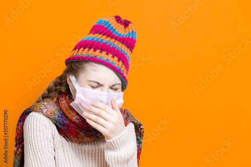 Healthcare Ideas.Depressed Teenage Female Girl in Warm Knitted Hat and Scarf Using Facial Mask Against Viruses While Coughing Over Trendy Yellow Background. © danmorgan12