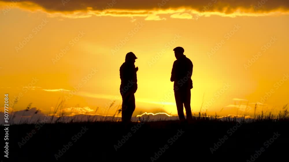 Two silhouettes of men talking at sunset or sunrise with dramatic sky and clouds.Dialogue and meeting two people on the horizon and skyline.Outlines of people in the sunlight.Golden hour with friends Two silhouettes of men talking at sunset or sunrise with dramatic sky and clouds.Dialogue and meeting two people on the horizon and skyline.Outlines of people in the sunlight.Golden hour with friends