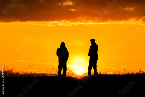 Two silhouettes of men talking at sunset or sunrise with dramatic sky and clouds.Dialogue and meeting two people on the horizon and skyline.Outlines of people in the sunlight.Golden hour with friends