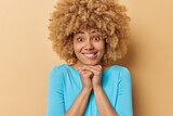 Positive young woman with curly hair bites lips keeps hands under chin dressed in casual blue jumper focused directly at camera isolated over beige background. Human face expressions concept