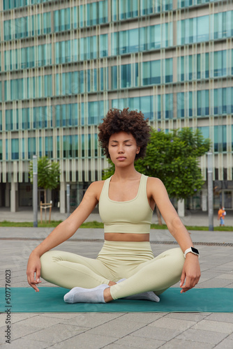 Photo of relaxed sporty young woman sits in lotus pose on fitness mat wears cropped top and leggings against cityscraper tries to relax breathes deeply. People meditation and healthy lifestyle