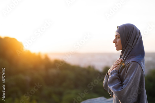 Young beautiful girl in a hijab prays and meditates on a background of sunset and landscape