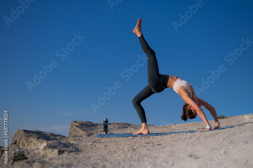 Beautiful slender caucasian girl in sportswear practices yoga on a rock on a background of blue sky