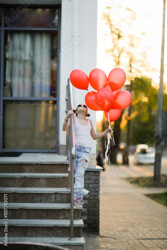 European child preteen girl in torn jeans with red balloons on stairs sunny summer day. Child in sunglasses at sunset on city streets, generation Z, happy birthday © natalialeb
