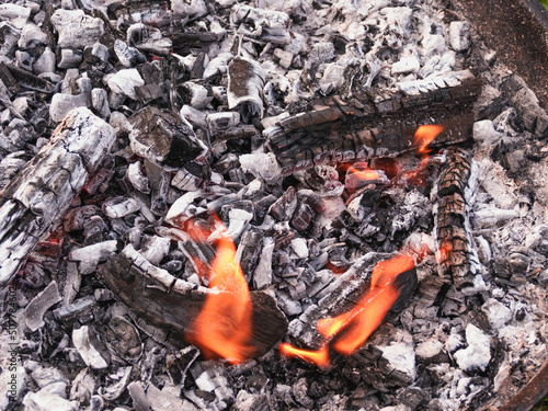 Dogasające ognisko. Warstwa żarzących się węgli pokrytych szarym nalotem popiołu. Spomiędzy węgli wydostają się pojedyncze, niewielkie płomienie.