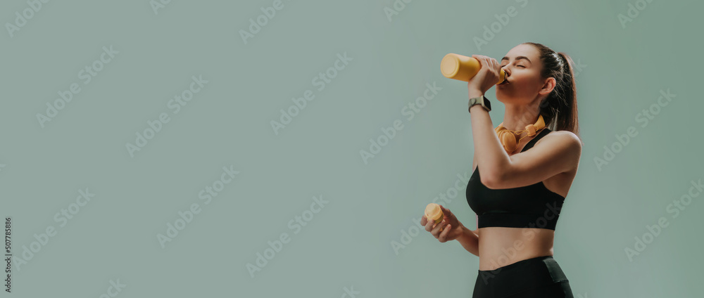 Young sporty woman in sportswear with wireless headset drinking from bottle over green background, copy space - obrazy, fototapety, plakaty 