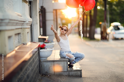 Funny stylish European girl child with red balloons in torn jeans sits on stairs on sunny summer day. Child in sunglasses at sunset on city streets generation Z, happy birthday