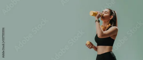 Obraz Young sporty woman in sportswear with wireless headset drinking from bottle over green background, copy space